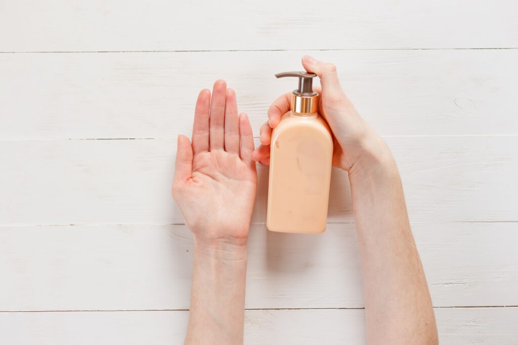 Is Hand Cream the Same as Lotion? What Dermatologist Says!!! Close-up of a person's hands applying lotion using a pump bottle on a white wooden background.