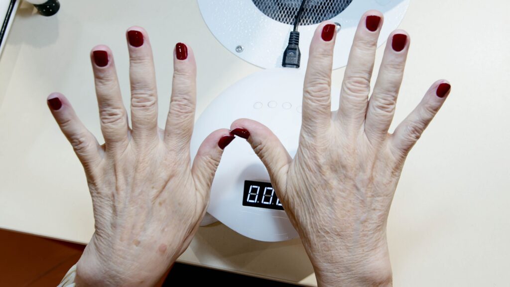 What Does Dry Hand Look Like? Close-up of senior woman's hands with red nails under drying lamp in salon.