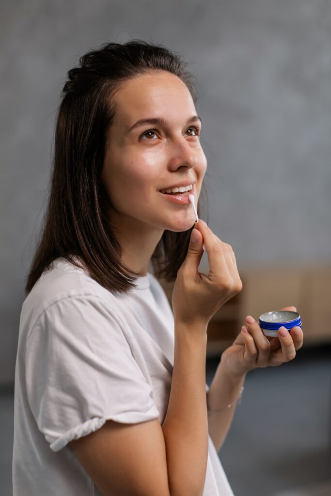 Is Vaseline Good For Dry Hands (Petroleum Jelly) Woman in a white shirt applies lip balm with a smile, showcasing a skincare routine.