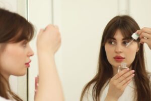Is PDRN Skincare Safe for Acne-Prone Skin? A young woman applying skincare serum in front of a mirror indoors. Perfect for beauty and self-care themes.
