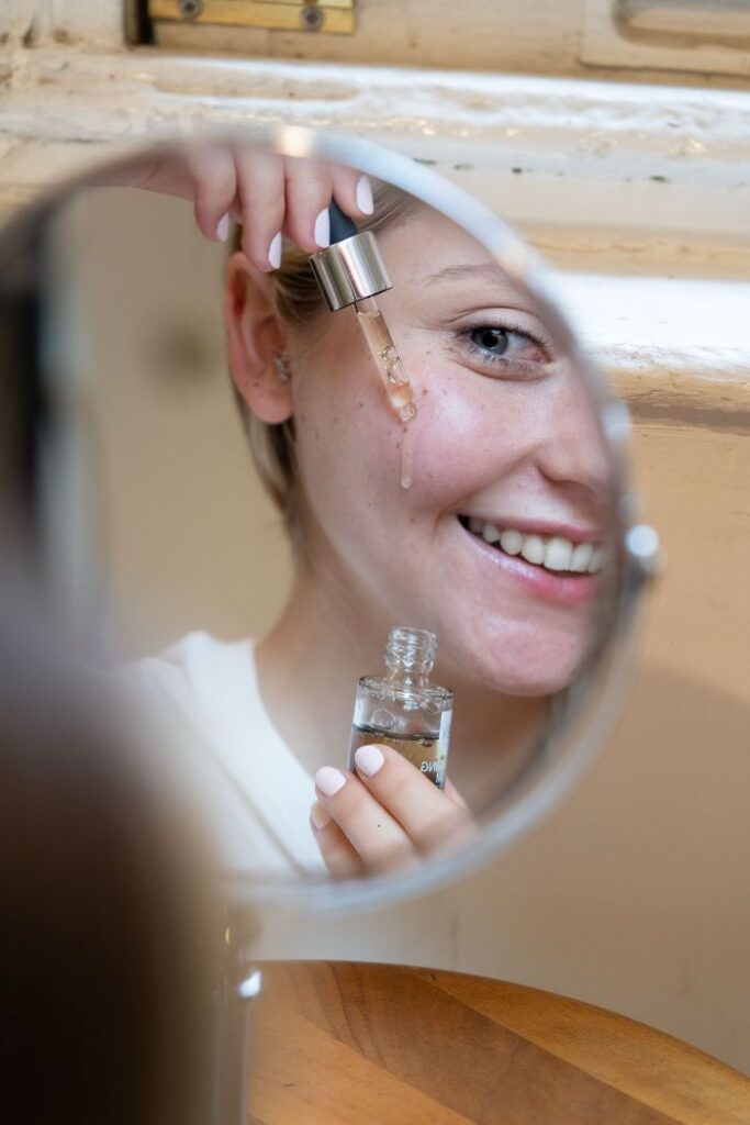 pexels photo 29745246 29745246 Smiling woman applying face serum with dropper seen in mirror reflection.