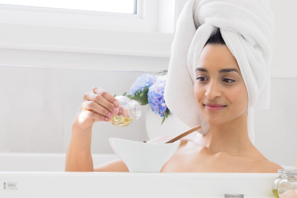 How to Do Oil Cleansing With Coconut Oil Step by Step A woman with a towel on her head adding essential oils to a bath in a bright bathroom.