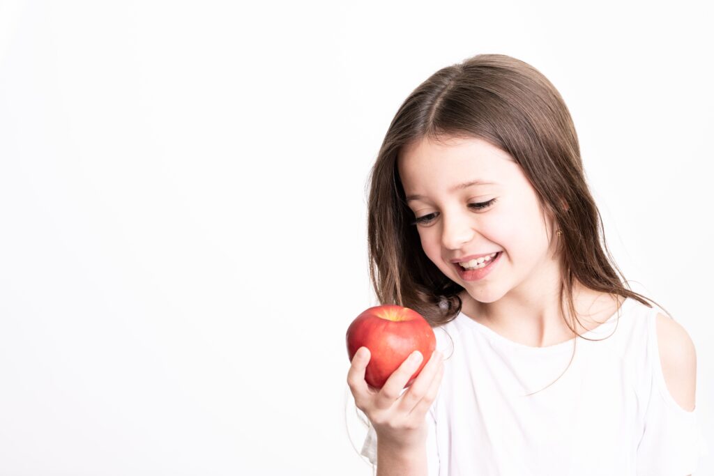 Positive young girl with brown hair in white clothes holding red apple and looking at it on white background