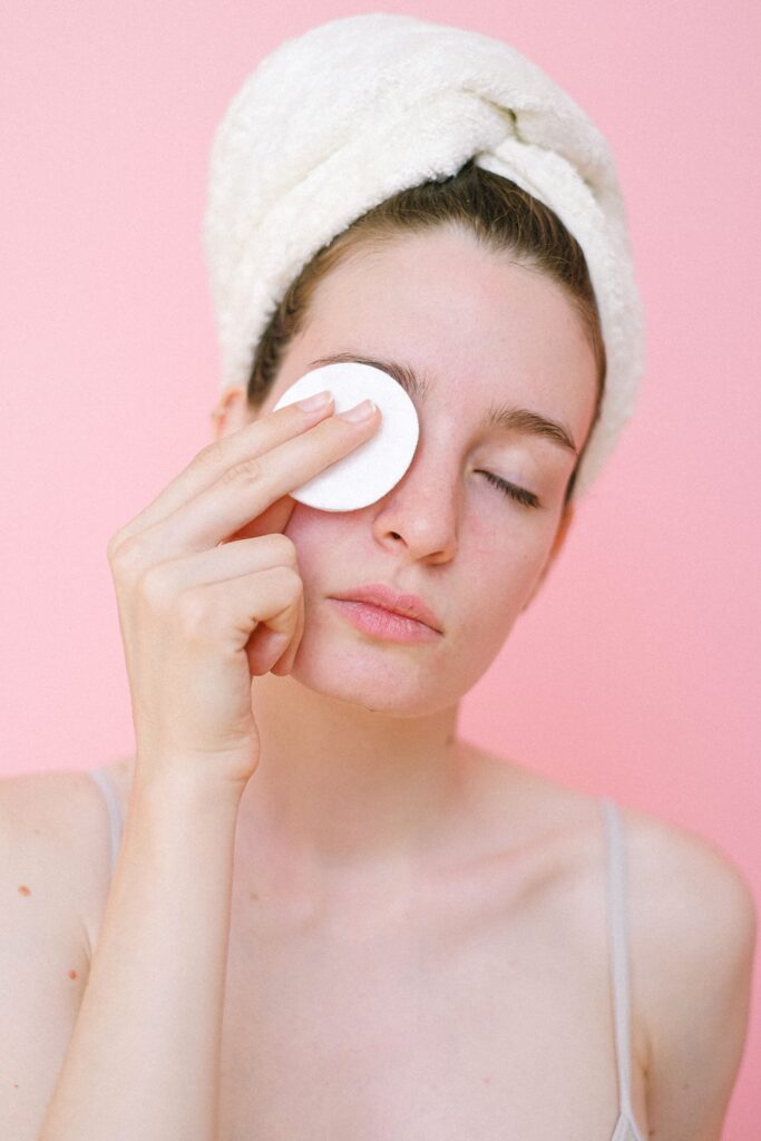 Young pretty female with perfect skin with hair wrapped in towel removing eyes makeup with cotton pad in light bathroom