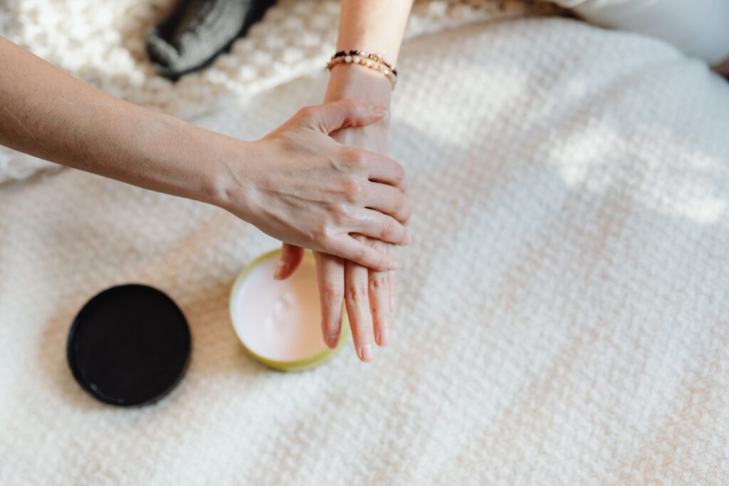 What is the Best Hand Cream for Dry Hands? A close-up of a woman applying moisturizer on her hands with a cream container on a cozy bed.