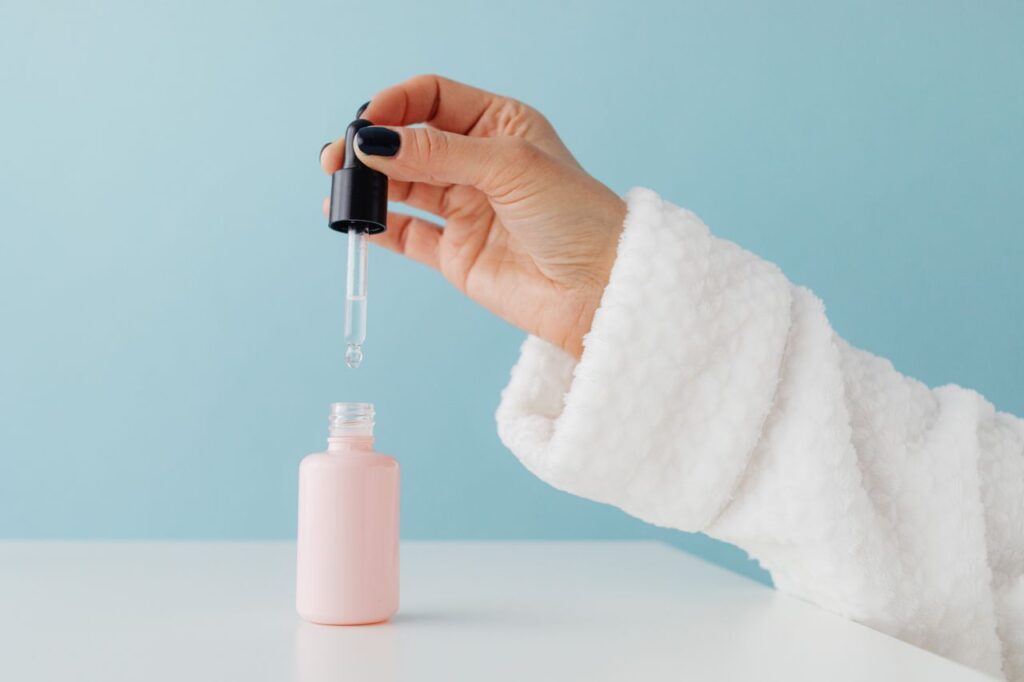Can Hyaluronic Acid Cause Breakouts? Close-up of a woman's hand in a bathrobe using a serum dropper over a pink glass bottle against a blue background.