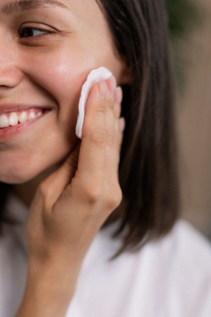 How to Do Oil Cleansing With Coconut Oil Step by Step Close-up of a smiling woman using a cotton pad on her face for skincare routine indoors.