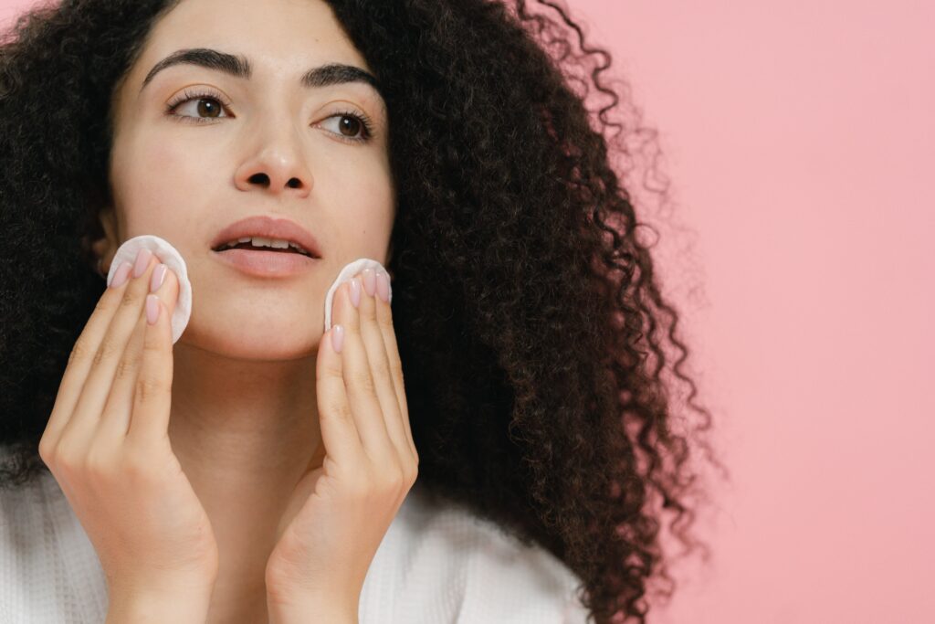 Close-up of a woman cleansing her face with cotton pads against a pink background.