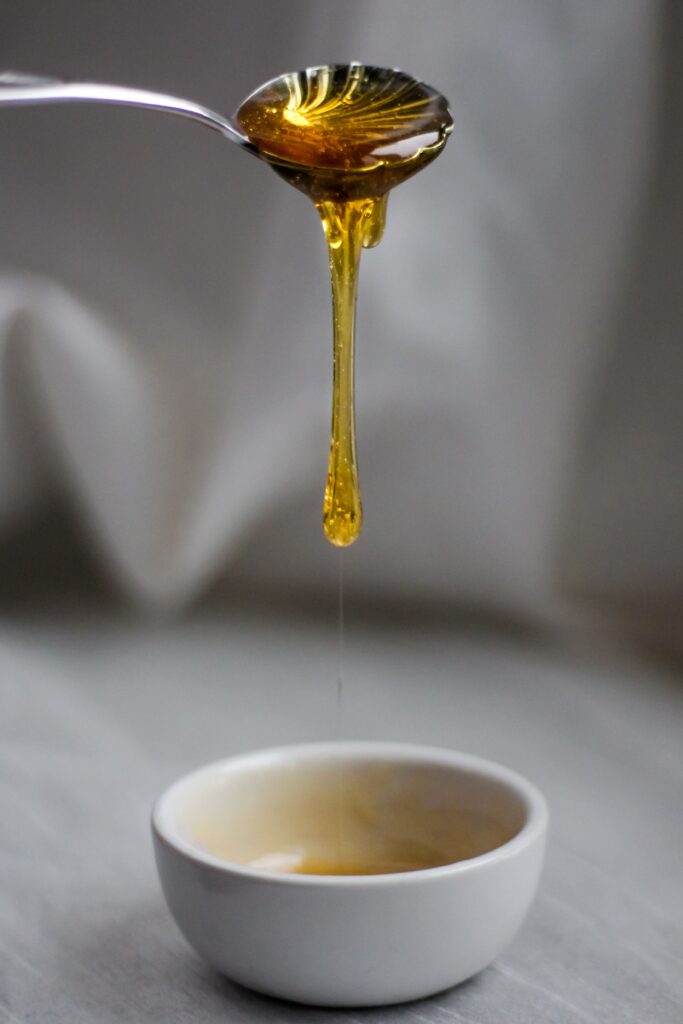 How to Brighten Your Skin Naturally at Home? Golden honey dripping from a spoon into a bowl, captured in a close-up shot with soft lighting.