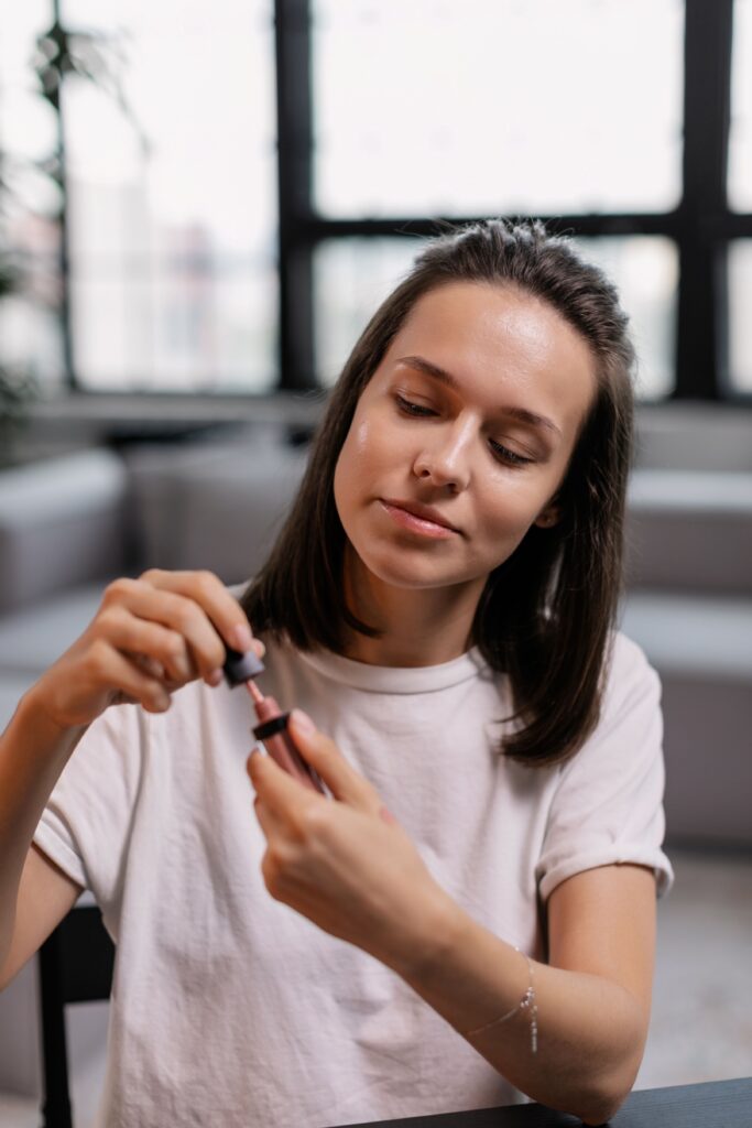 A woman in casual attire applying lipstick indoors with a thoughtful expression.